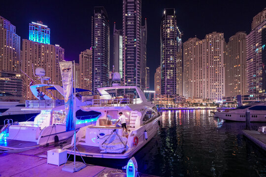 Dubai, United Arab Emirates - 22 April 2023: View of luxury yachts docked at Dubai Marina at night with a worker on deck and illuminated skyscrapers in the background.