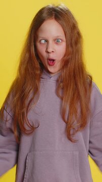 Cheerful funny bully young cute girl showing tongue making faces at camera, fooling around, joking, aping with silly face, teasing. Preteen redhead child kid on yellow background. Little children