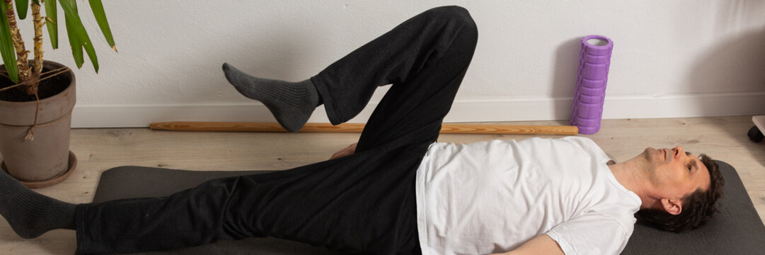 A 53-year-old man performs a leg stretch at home on a mat. He lies on his back with one leg raised and the other leg flat on the ground. He is focused on his workout routine, banner