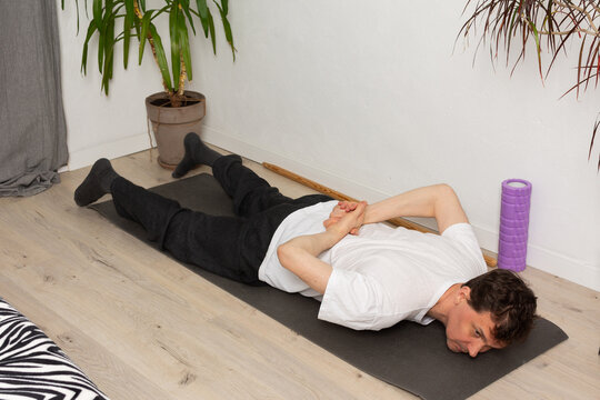 A man, 53 years old, lies on a yoga mat at home, performing a stretching exercise. He focuses on his fitness routine in a simple indoor space with plants around