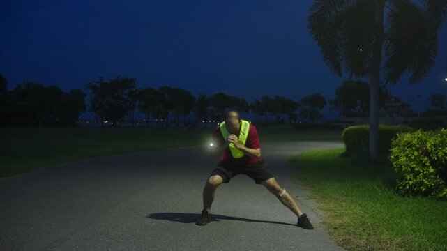 Middle-aged Asian man performing dynamic stretching before night jogging with LED safety light