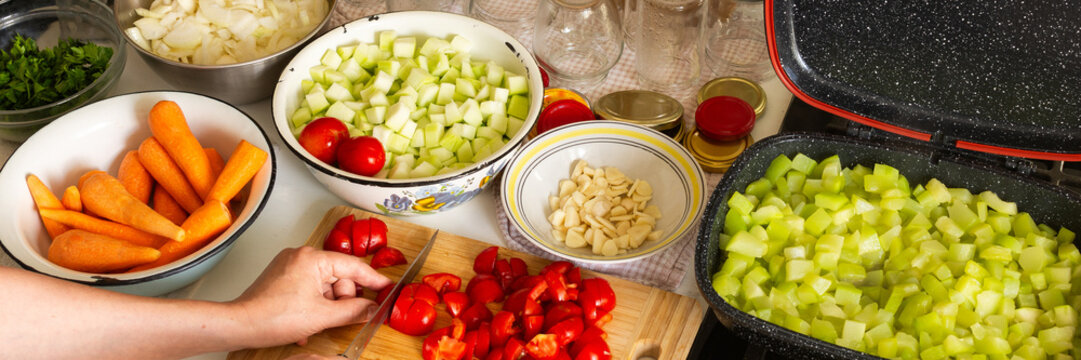 A woman cuts tomatoes on a wooden board surrounded by fresh vegetables while preparing squash caviar for winter canning in her kitchen, banner