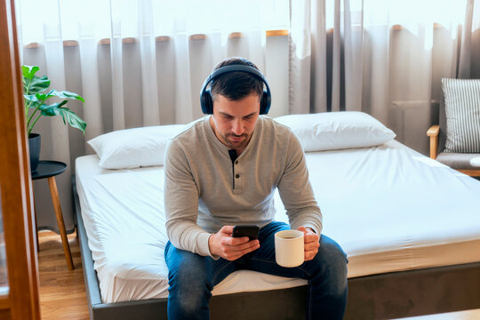 Caucasian young adult man sitting on bed, checking smartphone while wearing headphones and holding coffee, representing remote morning routine. Useful for work life balance or home tech ads