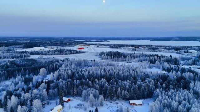Small town surrounded by frost covered forest, Lapland, Finland. Elevated drone view of red roofed buildings and frozen lake beyond hoarfrost-laden conifers under full moon in pale blue sky.