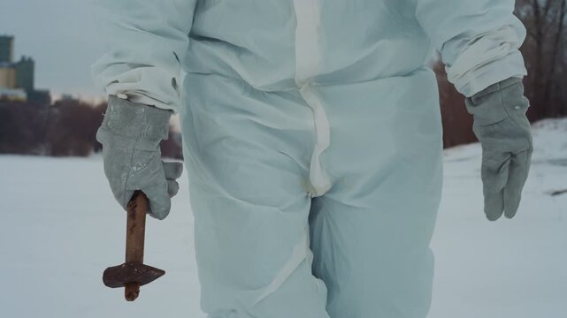 Hazmat suited scientist walking across snow holding rusted crucifix, gloved hand close up, abandoned riverbank and distant industrial skyline, cold pale light and eerie silence, slow deliberate steps