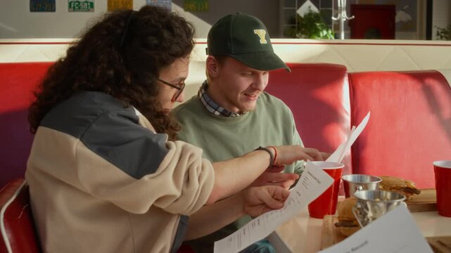 Medium shot of two diverse young male friends discussing academic records while hanging out at fast food cafe, comparing exam grades, arguing, among hamburgers, fries and soda on table