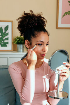 Vertical shot of young adult woman applying makeup with brush while looking into mirror at home, preparing beauty routine. Ideal for skincare, cosmetics, self care, lifestyle marketing