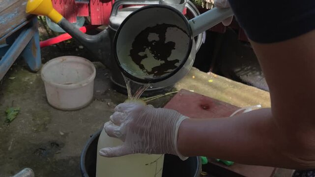 Unrecognizable gardener wearing rubber gloves pouring liquid fertilizer from metal dipper into hand pump pressure sprayer outdoors on sunny summer day in rustic garden close up shot authentic footage