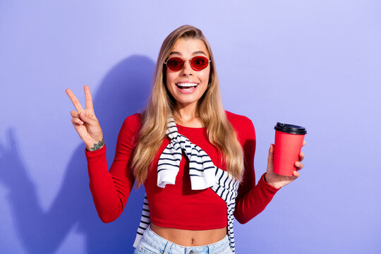 Smiling young woman in red top holding takeaway coffee and flashing peace sign with sunglasses for casual lifestyle portrait