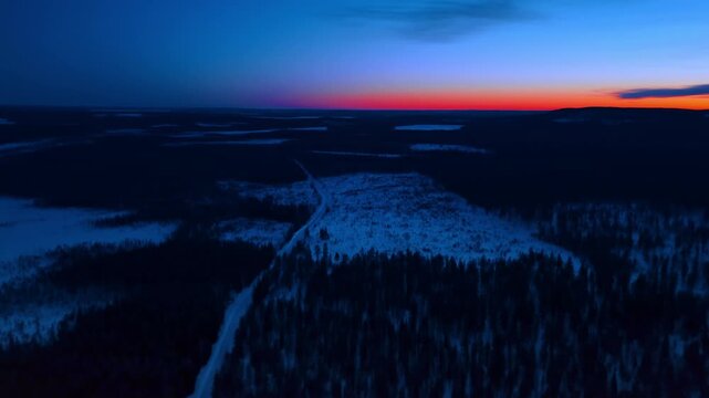 Snow road through arctic forest under vivid red sunset, Lapland, Finland. Narrow winter road cutting through dark conifers, blue snow fields and fiery orange red horizon at nightfall.