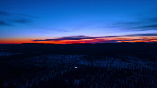 Polar night sunset glow over dark boreal forest, Lapland, Finland. Drone panorama of silhouetted taiga under vivid red and orange horizon band fading to deep blue sky.