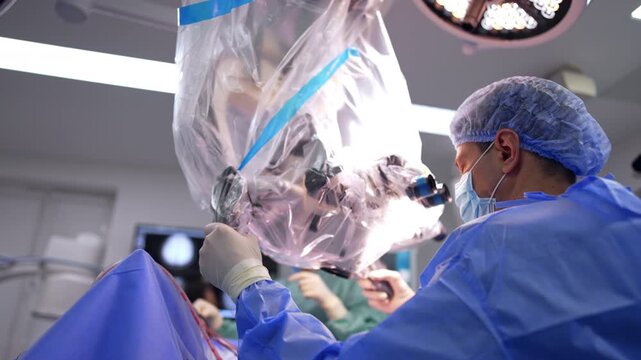 Neurosurgeon using draped surgical microscope during brain operation. Surgeon at eyepiece with gloved hands on drape, team member and monitor visible behind.