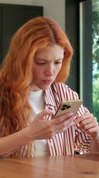 Young frustrated woman with long red hair reacting to bad news on her phone, showing expressions of sadness, disappointment, anger, and stress while sitting at a table in her home