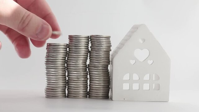 A hand reaches out and puts coins in a stack near a small white ceramic house on a white table. The stack grows noticeably as coins are added one after another,