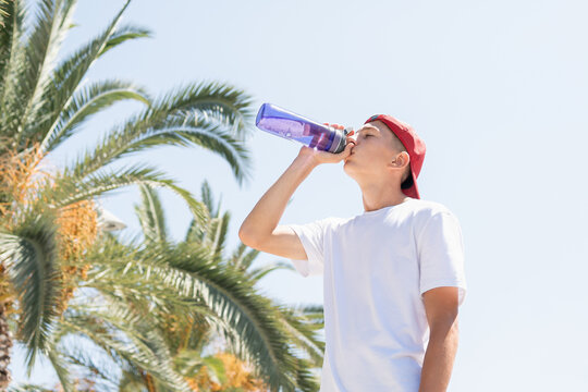 Teenager boy drinking water outdoors under palm trees on a hot summer day. Healthy hydration lifestyle, reusable bottle and tropical vacation mood in bright sunlight