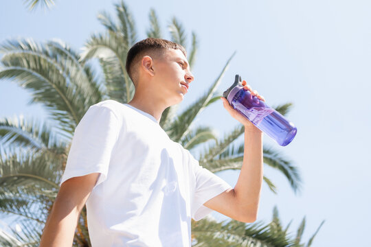 Teenage boy enjoying cold water outdoors in tropical city setting. Gen Z summer lifestyle concept with reusable bottle, sunny weather and healthy daily routine. Travel, summer heat, hydration