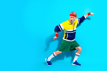 Obraz premium Senior man in retro tracksuit exercising and striking a dynamic pose on bright blue background — active fitness, playful energy and colorful sportswear for mature lifestyle and wellness