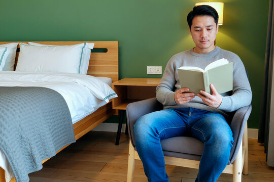 Asian young adult man reading book while sitting in bedroom, studying and relaxing during solo stay. Useful for hospitality, self care, leisure, lifestyle marketing