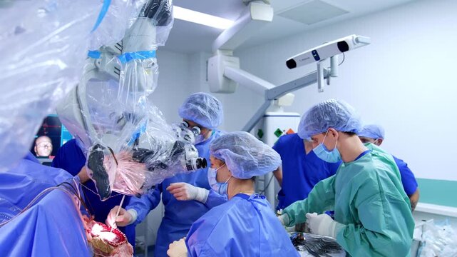 Surgical team using microscope during open neurosurgery with neuronavigation camera above. Multiple surgeons in blue and teal gowns around lit operative field.