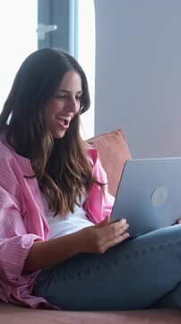 Excited young woman sitting on a sofa receiving amazing news on her laptop, celebrating her success with genuine emotion, feeling euphoric and happy about her online achievement at home