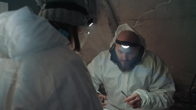 Caucasian researcher in protective suit contemplates contaminated vial while seated in ruined shelter under dim headlamp, bearded colleague watching, breath misting in cold, somber reflective mood