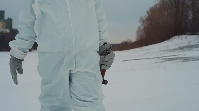 Scientist in protective suit holds metal cross near frozen field, closeup of gloved hand gripping rusted artifact, bleak snowplain, muted sky, abandoned perimeter suggesting nuclear fallout and tense