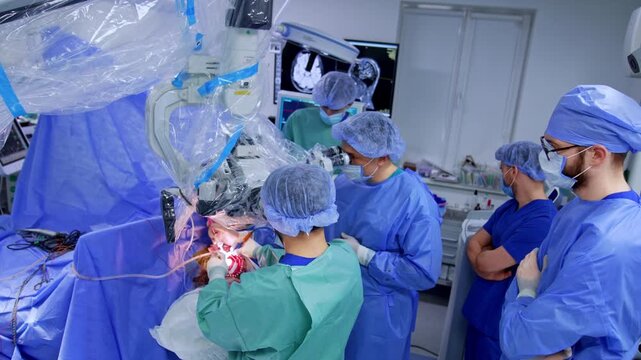 Neurosurgical team at operative field with brain CT on monitor behind. Surgeons in blue and teal gowns, draped microscope, observers watching at right.