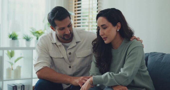 Young Hispanic husband comforting sad depressed wife on sofa in home living room. Mental health support, marriage couple conflict problem, grief emotion therapy, family trauma crisis concepts.