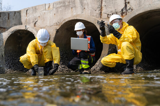 Environmental scientists in hazmat suits and safety gear taking water samples for toxic chemical pollution testing near a sewage drainage pipe with a coworker using laptop