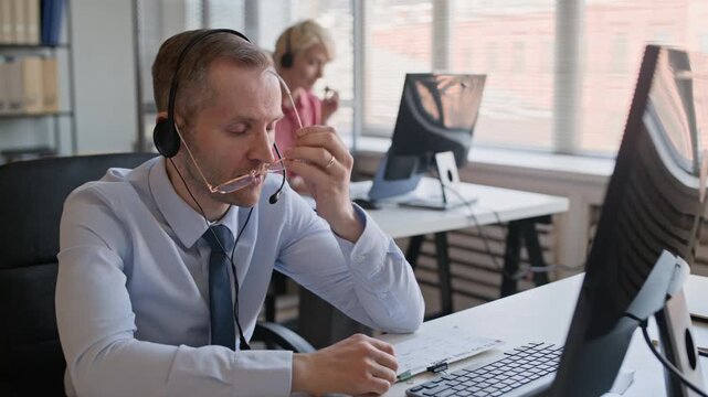 Medium shot of patient male support agent taking off glasses while repeating instruction to client via headset sitting at desk in modern call center