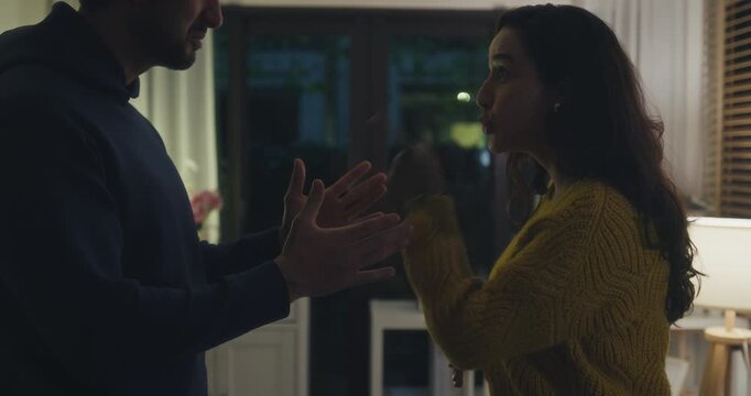 Angry Hispanic young couple arguing and pointing fingers in dark home living room at night. Domestic conflict dispute, toxic relationship problem, divorce crisis, communication breakdown concept.