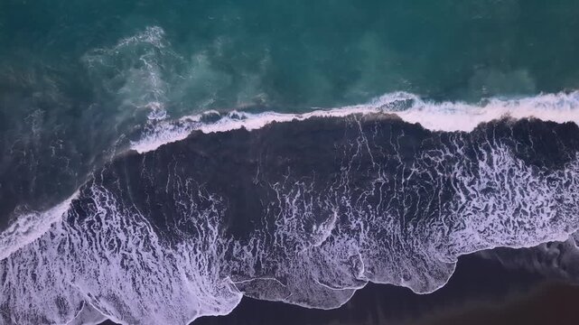 Top down aerial view of turquoise ocean waves crashing on a black sand beach