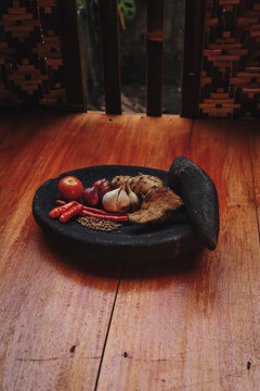 various traditional Indonesian cooking spices arranged inside a black stone mortar (cobek) with its pestle (ulekan) resting on the side.