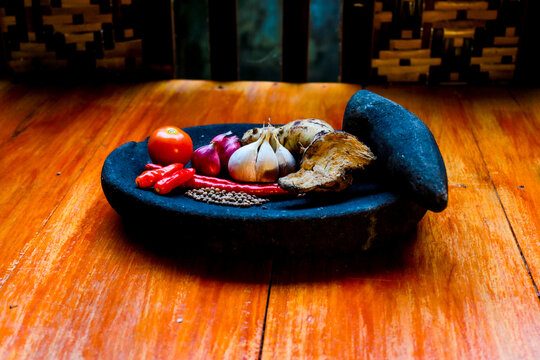 various traditional Indonesian cooking spices arranged inside a black stone mortar (cobek) with its pestle (ulekan) resting on the side.