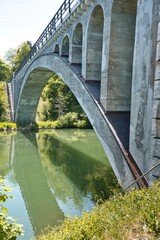 Fototapeta premium Historic concrete arch bridge over a calm green river in a lush forest landscape.