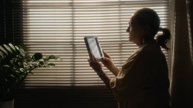 Lowkey medium shot of older woman standing near window with closed blinds and looking at framed portrait in warm apartment interior