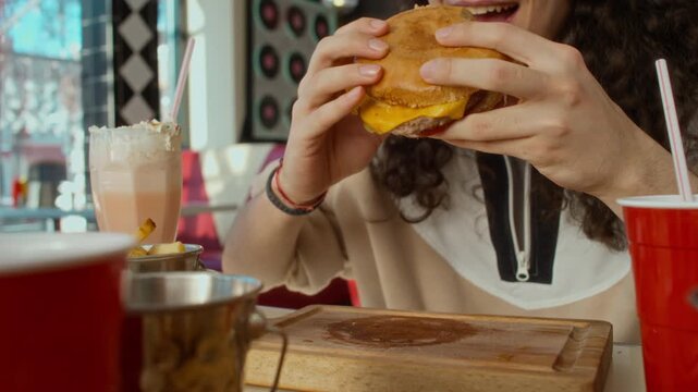 Medium close-up of lower face of eager young gen Z Caucasian man sitting at table in fast food restaurant, rubbing hands in anticipation, then tucking into tasty hamburger and chewing with pleasure