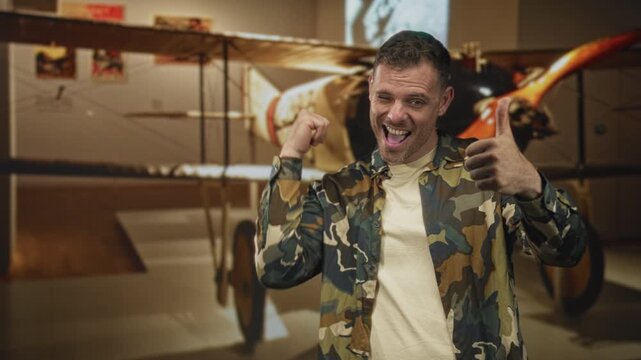 Young hispanic soldier in camouflage jacket gives thumbs up smiling in front of vintage biplane inside building; pride duty.