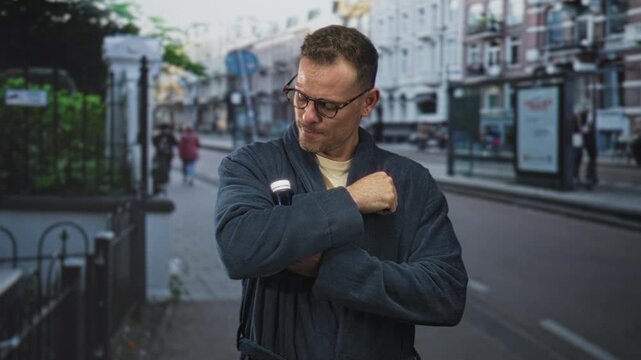 Man in robe and glasses holds a pill bottle to chest with hand across chest on a street lined with buildings; relief recovery.