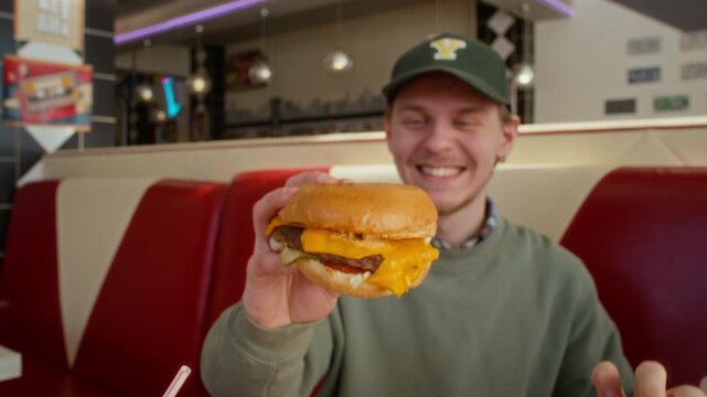 Medium slowmo shot of young Caucasian man enjoying fast food meal together at restaurant with group of friends, toasting each other with tasty gourmet cheeseburgers while laughing