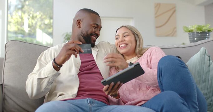 Mid-adult couple at home on sofa, scrolling tablet while man raising credit card preparing payment