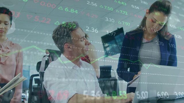 Lead pointing at laptop urging standing woman writing notes while financial tickers scrolling