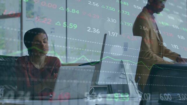 Seated analyst typing on laptop starting stock overlay while coworker passing for market review