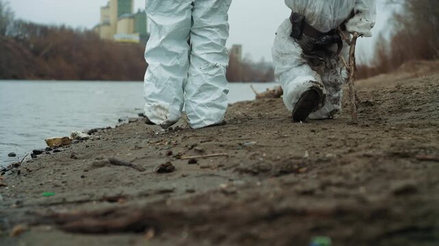 White scientist father and child riverside, wearing soiled protective suits, trudging through mud and debris, leaving heavy footprints beside polluted water, industrial plant looming in distance, cold