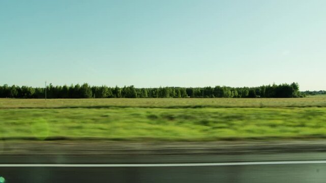 Long shot of blur motion across green field from vehicle window. Landscape shows forest on distant horizon and wide blue sky, capturing essence of road trip during sunny day.