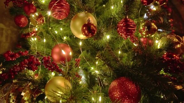 Close up view of decorated Christmas tree. Numerous glowing warm white string lights illuminate textured red and shimmering gold sphere ornaments. Artificial pine branches provide festive holiday back