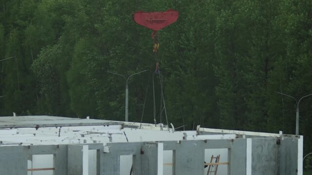Large red crane hook lowers concrete slab onto partially built structure. Concrete wall panels form foundation with window openings. Green forest and street lights visible.