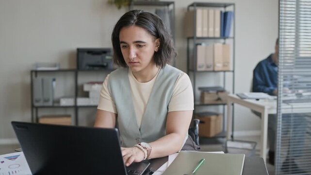 Medium portrait of confident female Caucasian employee with disability doing report on laptop and looking at camera in modern office