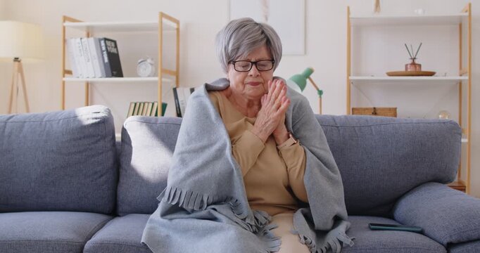 Old grey haired woman wrapped in plaid as feeling cold, trying to warm herself, has problems with heating at home, sitting on sofa in living room during winter season. Power crisis concept
