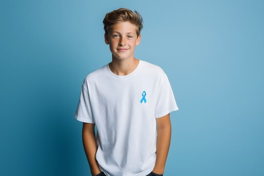 Portrait of a teenage boy 16-18 years old wearing white t-shirt with blue awareness ribbon pinned on chest against blue background. Youth-focused health awareness image for teenage men's health campai